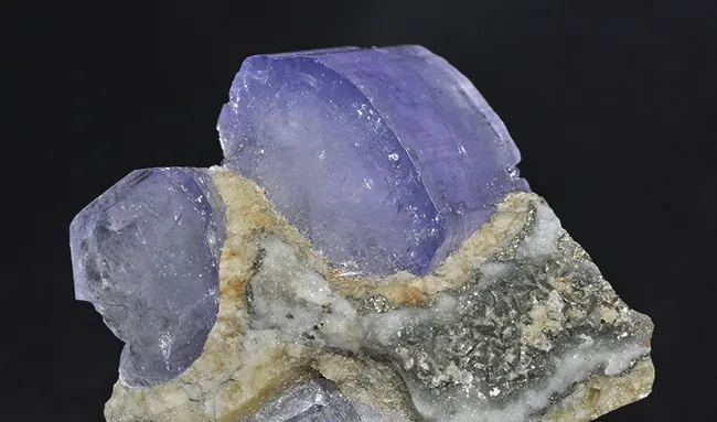 Esta figura incluye una imagen de dos cristales grandes de apatita de color azul claro en un conglomerado mineral que incluye cristales blancos, grises y bronceados. Los cristales de apatita azul tienen un aspecto apagado, polvoriento o pulverizado.
