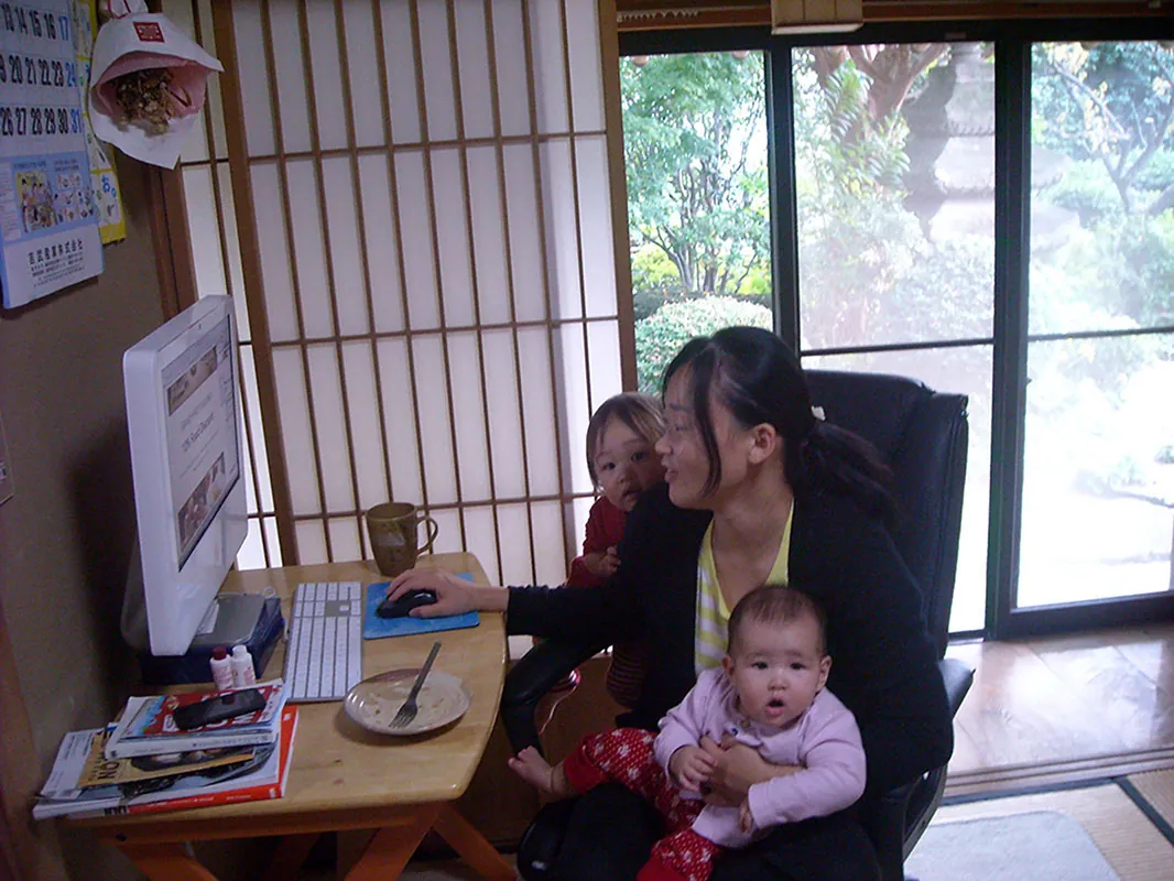 A person sits at a desk while working at a computer, while holding a baby of about six months old. A second child leans against the chair as well.