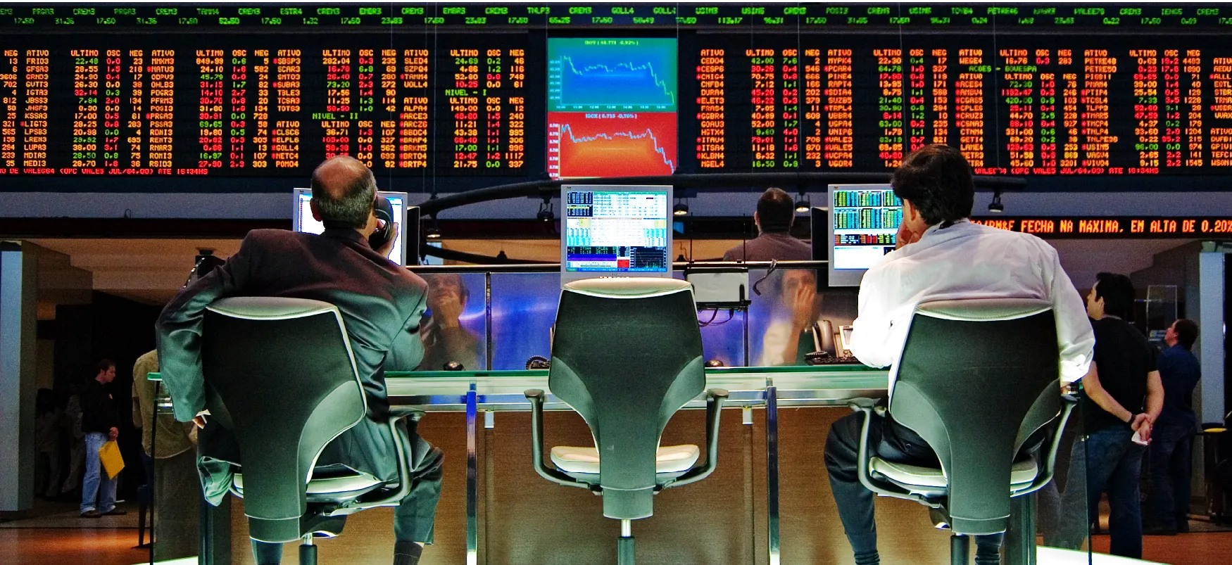 Two stockbrokers sit on chairs and look at the stock tickers on a giant screen. Two graphs are displayed in the middle of the screen, showing a downward trend.