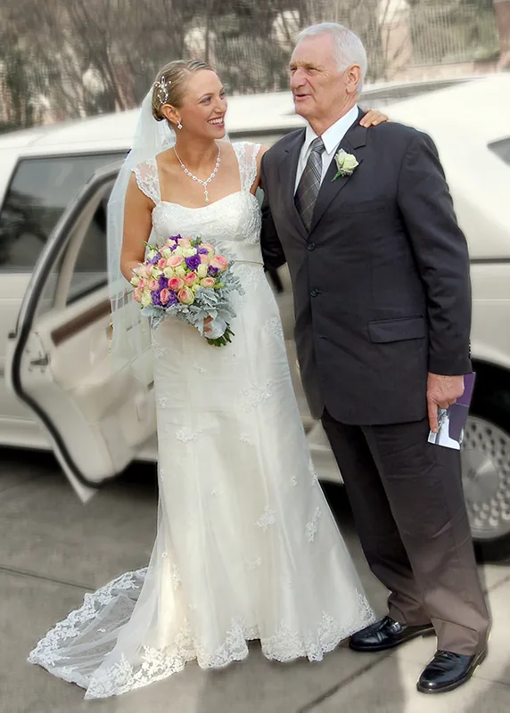 A bride wearing a long white wedding dress, is standing next to a mature man dressed in ta suit. Both are standing in front of a white limo.