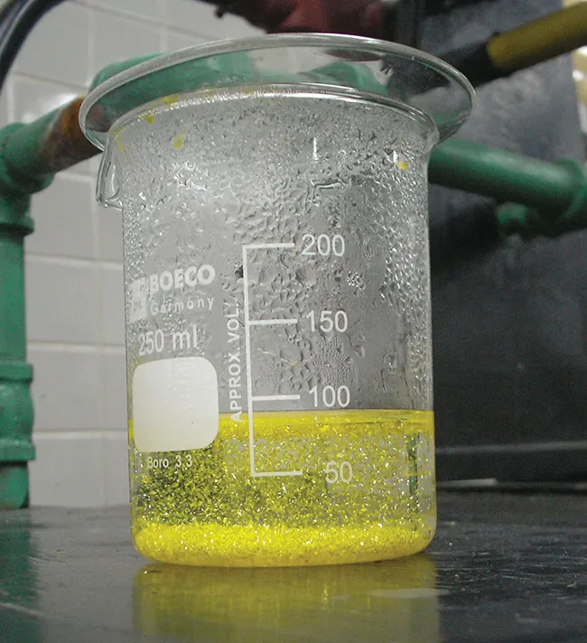 Se muestra una fotografía de una sustancia opaca de color amarillo verdoso agitada por un líquido claro e incoloro en un tubo de ensayo.