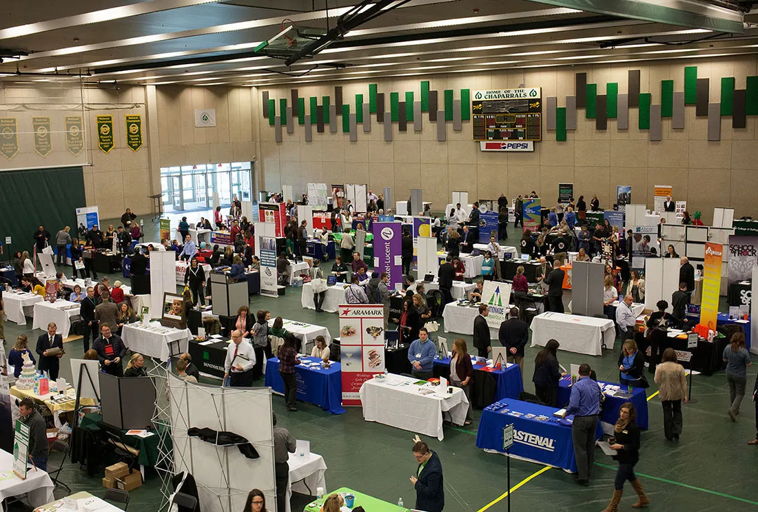 People attend a job fair. Several rows of tables are set up in a college gymnasium, and people walk between them and speak to each other.