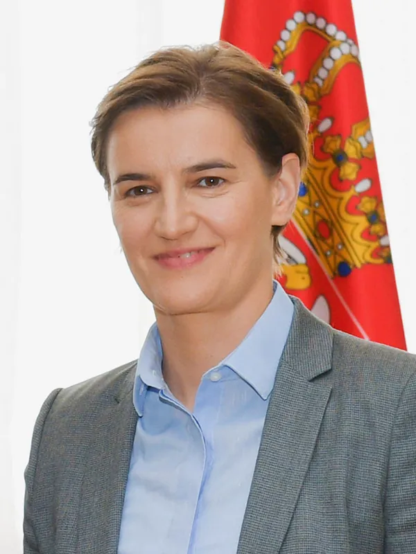 Serbian Prime Minister Ana Brnabić poses for a portrait in front of a bright red flag emblazoned with an image of a jeweled crown.