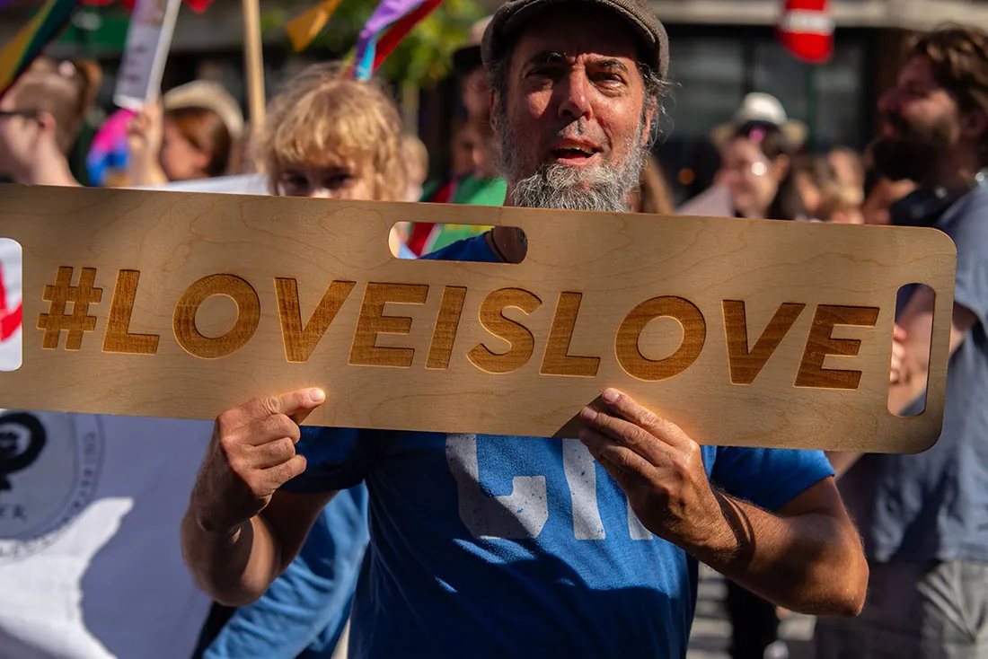 A person holds a sign at a parade or rally. The sign has a hashtag and reads Love Is Love.