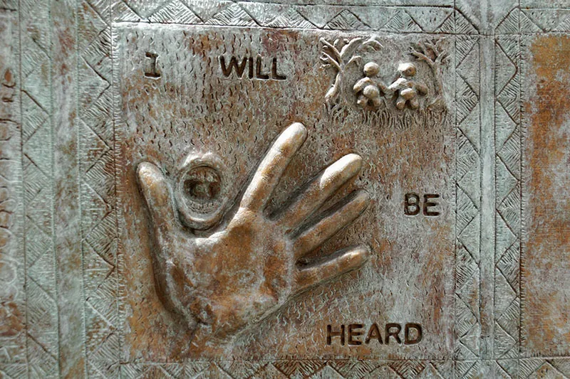 A relief sculpture contains a three-dimensional hand and mouth with the words “I Will Be Heard.”