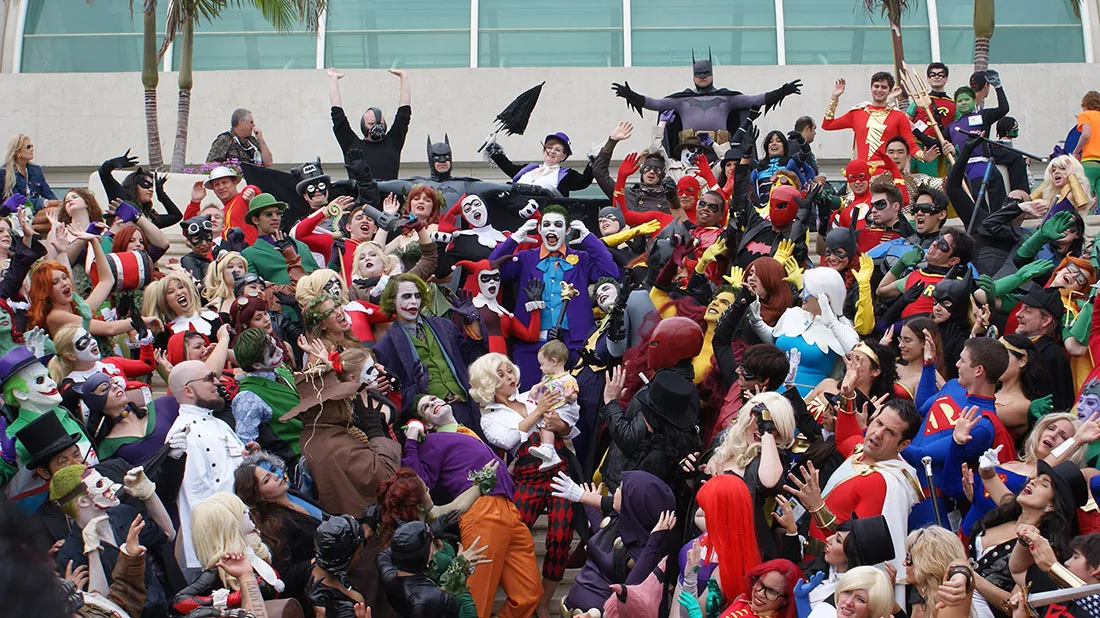 At least a hundred people wearing costumes stand in a rough circle around a central figure holding a baby. The costumes include Wonder Woman, Batman, Superman, Harley Quinn, the Joker, Mera, and others.