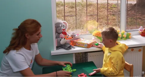 An adult and a child sit and talk at a child's table. Stuffed animals are on a shelf between them.