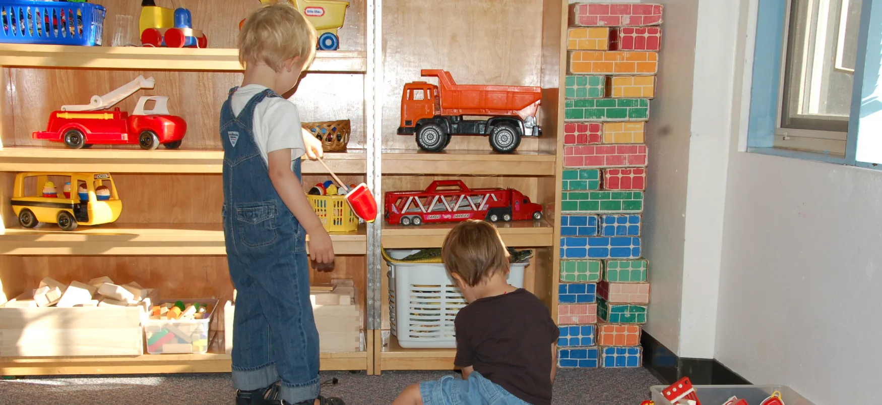 Two children are in front of shelves that are filled with toys such as trucks, buses, and building bricks.