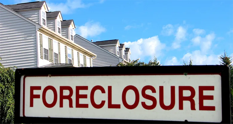 The image shows a “Foreclosure” sign in the foreground and the tops of a couple of houses in the background.