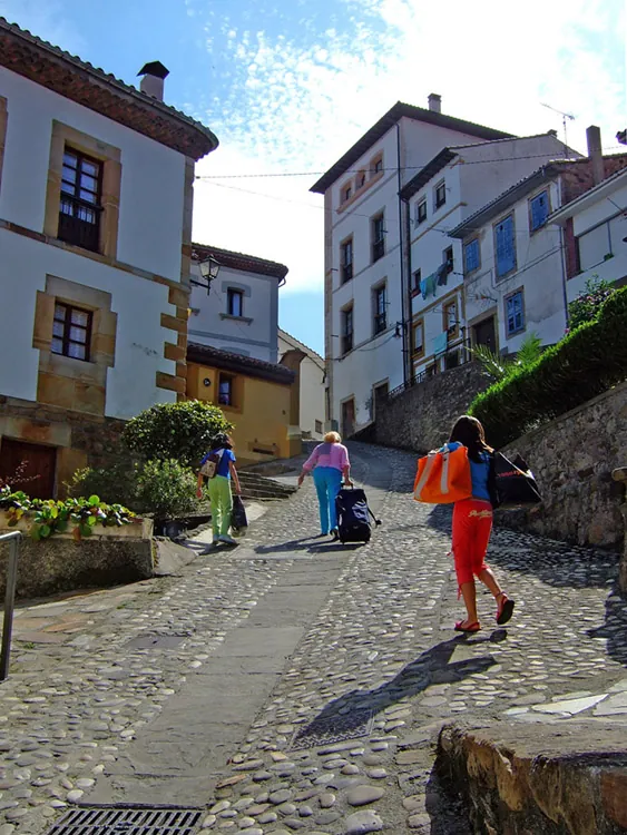 Three female tourists carrying luggage are shown climbing a cobblestone hill.