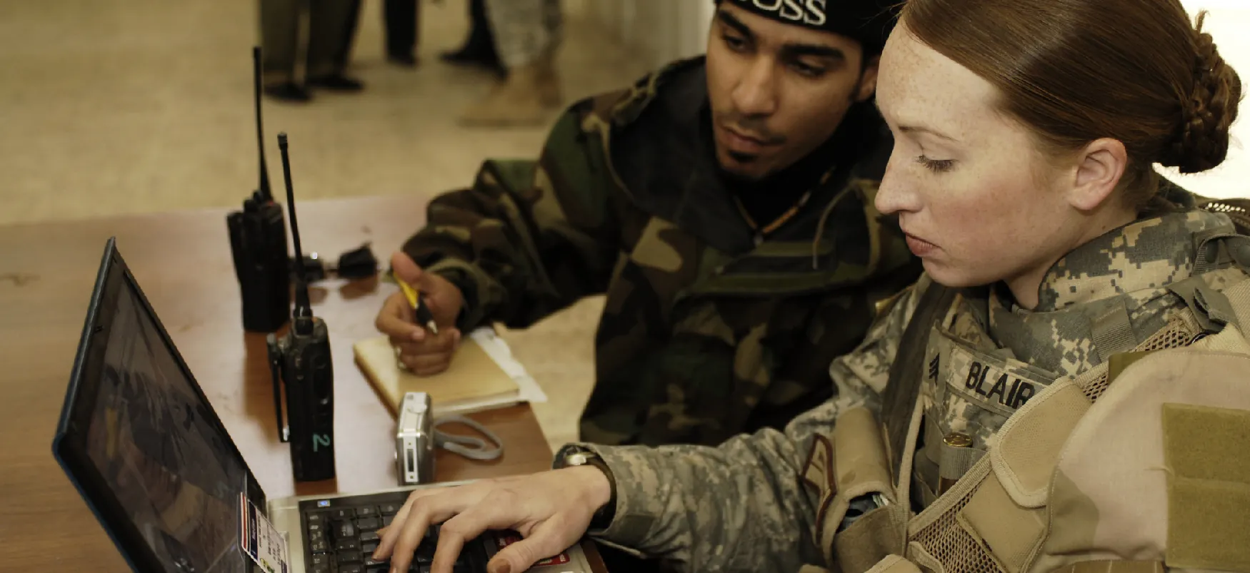 A military sergeant sits with an Iraqi police officer and enters the Iraqi police officer’s statements into a computer.