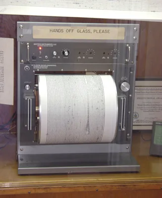 The figure shows a seismograph put on a wooden table. Its top is labeled as “Hands off glass, please”. Below it there are some buttons are shown and a paper roller is fitted in the seismograph to print the observation by the machine. On the right and left of the roller, two vertical cable slots are given.
