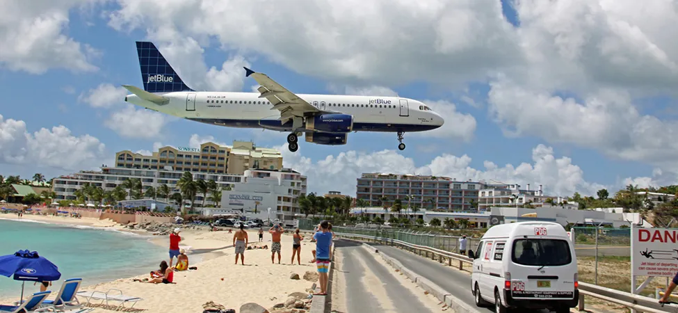 An airplane is flying over a beach area in St. Maarten as it comes in for a landing at the airport. The airplane's acceleration is in the opposite direction of its velocity.