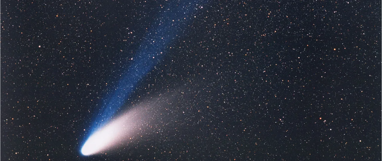 Comet Hale-Bopp. In this photograph the bright nucleus is seen at lower left, with the white dust tail curving away toward center-right. The blue ion tail moves away from the nucleus straight toward the top-center of the image.