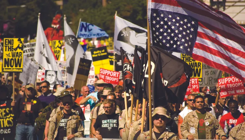 An image of a group of people, several of whom are holding flags and signs. One of the signs reads “End the war now”, and another reads “Support the troops, end the war”.