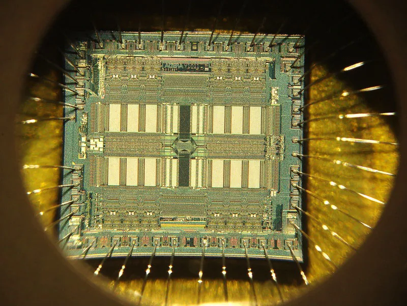 Magnified color photograph of a semiconductor chip. The magnification makes visible a complex pattern of shapes and lines across the surface of the chip.