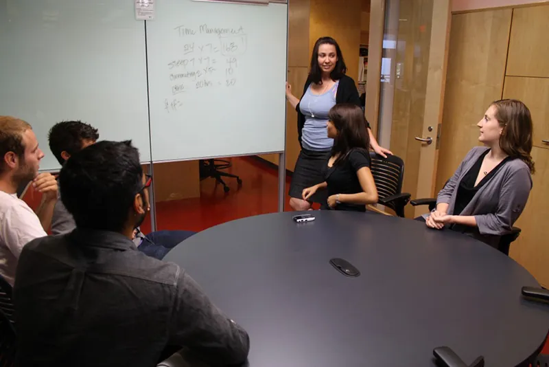 A group of five people sit around a table in a conference room. A sixth person is standing at the front of the room, next to a whiteboard with writing on it.