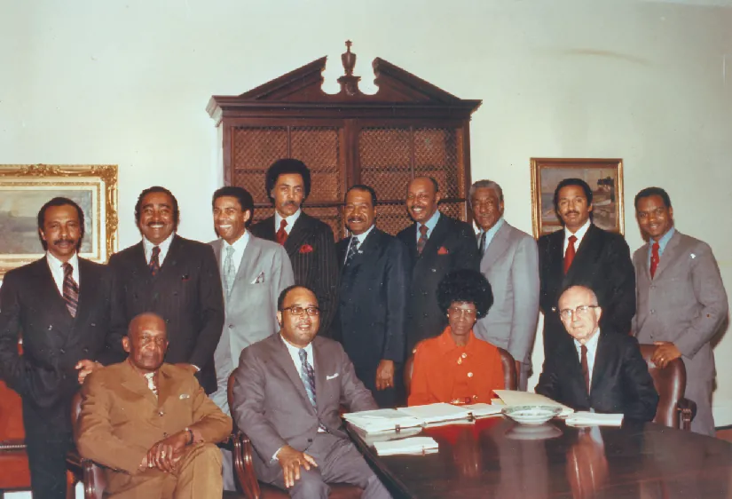 An image of a group of people, four of whom are seated at a table, and nine of whom are standing.