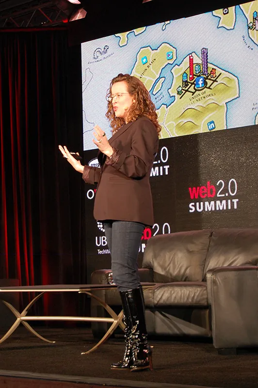 A woman with long curly hair and wearing a blazer, jeans, and boots stands on a stage speaking at a conference.