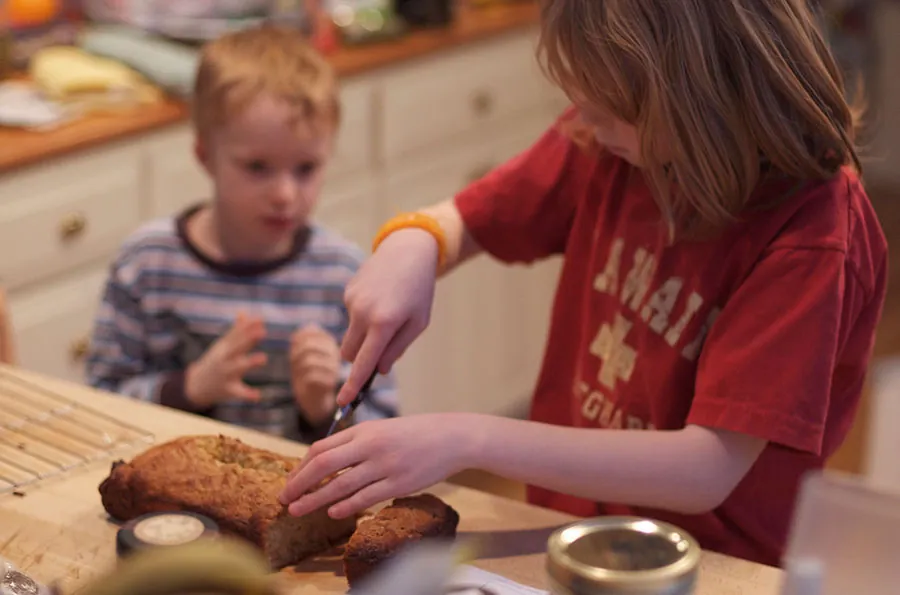 A child who appears to be about age eight cuts a loaf of bread while a younger child watches.