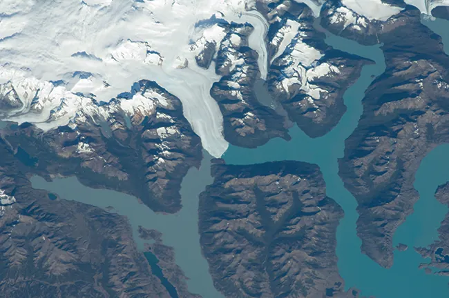 A photograph shows an aerial view of a land mass. The white mass of a glacier is shown near the top left quadrant of the photo and leads to two branching blue rivers. The open land is shown in brown.