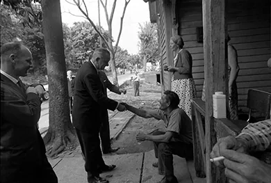 A photograph shows President Johnson standing on a street outside of a house, several of whose inhabitants sit and stand on the porch. He shakes the hand of a seated man while two other officials look on.