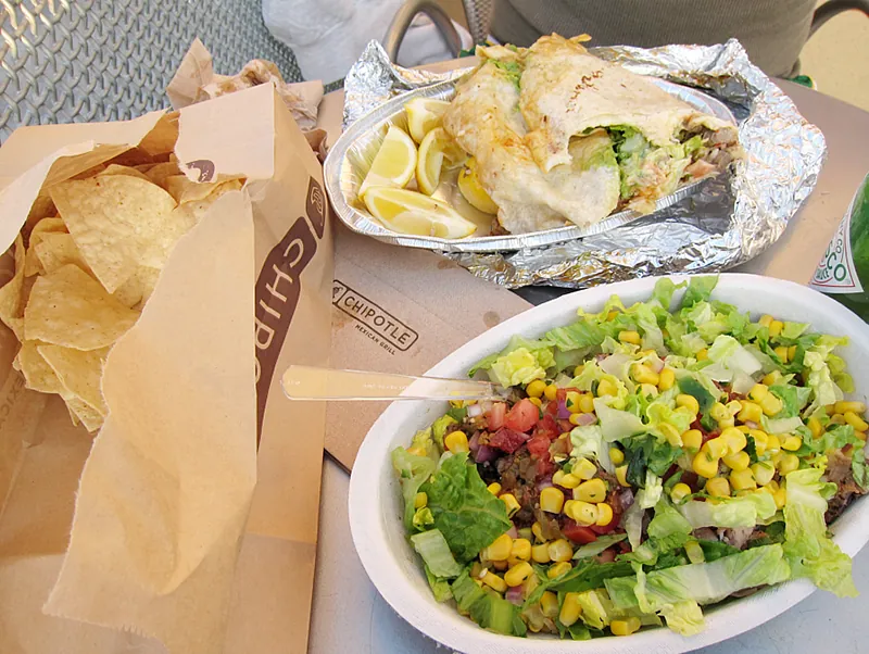 A meal from Chipotle is on a table. The tortilla chips are packaged in a brown paper bag while the burrito and carnitas bowl are in cardboard bowls. The tinfoil that the burrito had been wrapped in is off to the side.