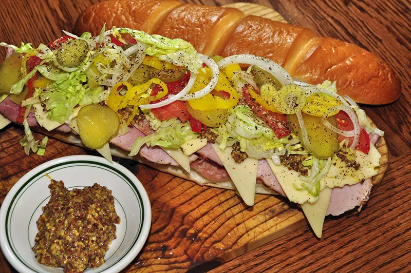 A submarine sandwich with lunchmeat, cheese, lettuce, pickles, tomatoes, and onions is on a table.