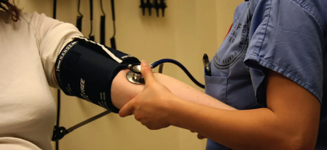 This photo shows a nurse taking a woman’s blood pressure with a blood pressure cuff. The nurse is pumping the cuff with her right hand and holding a stethoscope on the patient’s arm with her left hand.