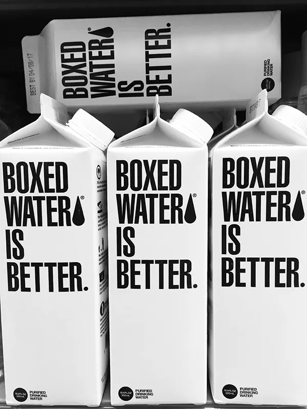 A photograph of four cartons of boxed water is better. Three boxes are next to each other in a row, while the fourth box  is lying horizontally on top. The boxed water is better logo takes up most of the space on each carton.
