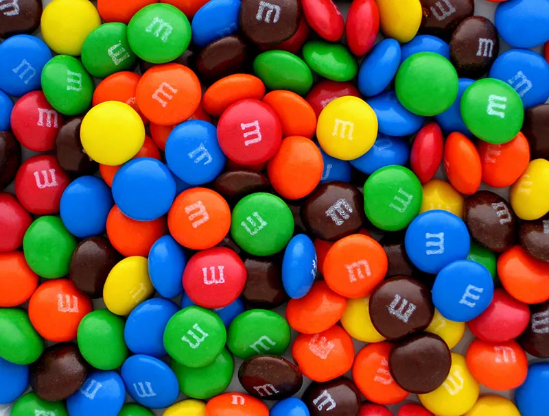 A mix of M&M candies of varying colors, red, blue, yellow, green, brown, and orange, feature the logo “m.”