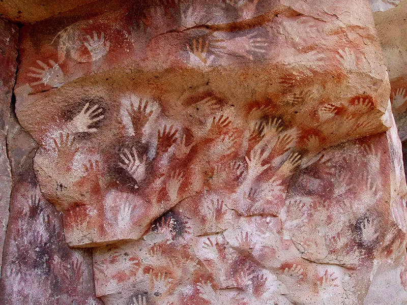 Cave wall decorated with dozens of overlapping hand prints in various colors and shades.