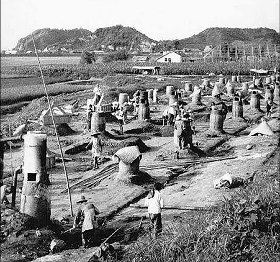 A photograph of a large field is shown with houses and hills in the background. In the foreground workers move about the field dressed in long shirts, pants, and large hats. There are many circular steel containers arranged in three rows in the field, some with square holes in them, some covered with pieces of cloth.