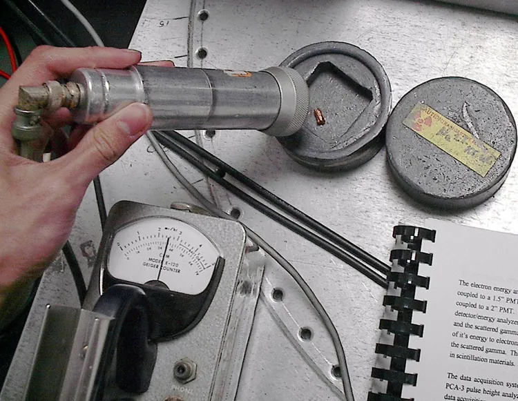 Image shows a person’s hand holding a cylindrical object placed near a small piece of radioactive material. A dial indicator is connected to the cylindrical radiation detector.
