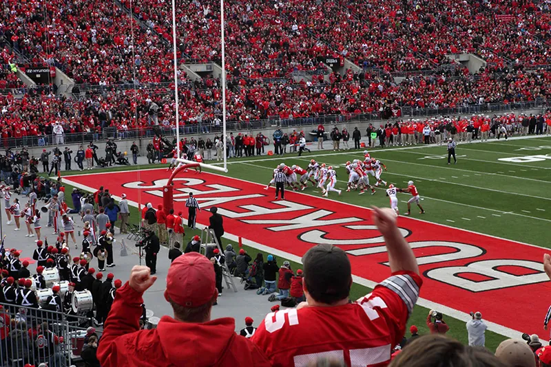 Many people are shown attending a football game. The football players are near the end zone.