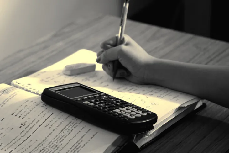 Una fotografía de la mano de un estudiante, quien trabaja en un problema con un libro de texto abierto, una calculadora y una goma de borrar.