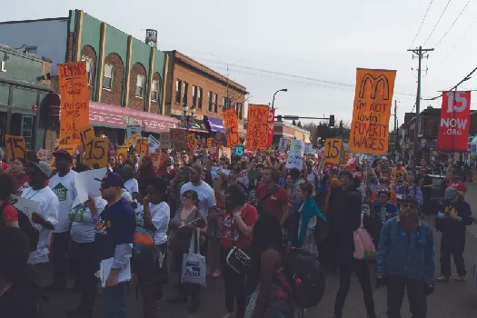 An image of a group of people marching down a street, one of whom holds a sign that reads “I’m not loving’ poverty wages”.