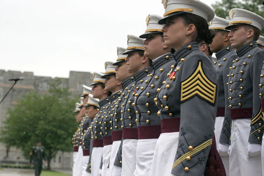 A group of West Point cadets in full dress gray uniforms with white peaked caps. Each wears a red sash around their waist, indicating that they are seniors
