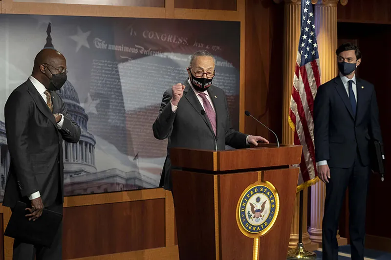 Georgia Senate candidates Raphael Warnock and Jon Ossof stand at either side of Chuck Schumer, who speaks before a podium emblazoned with the seal of the US Senate.