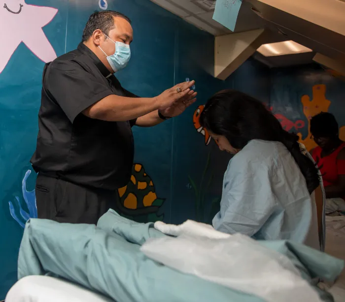A photograph shows a Catholic priest performing a blessing while holding his hands out over bent head of woman in hospital gown.