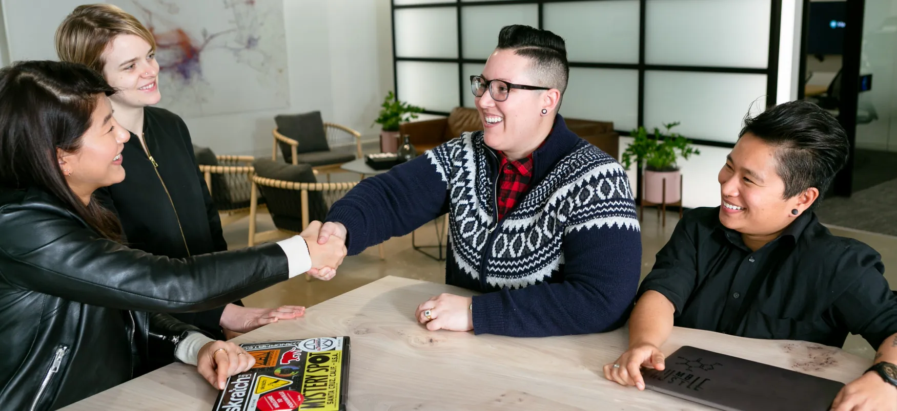 A group of four people stand around a table. Two of the people are shaking hands across the table.