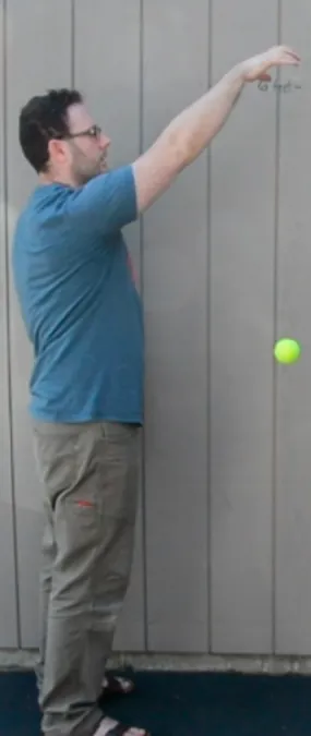 A man in a blue shirt and khaki pants stands sideways  against a gray paneled wall, arm extended upward, and has dropped a yellow tennis ball. The ball is approximately waist height of the man..