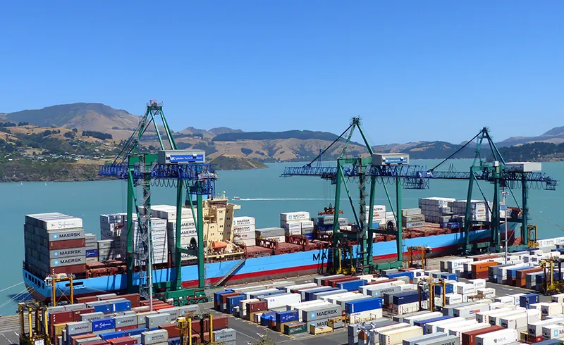 Tall cranes place shipping containers on a boat that is docked. Shipping containers are stacked on top of each other. Rows of shipping containers are also on the ground in beside the boat.