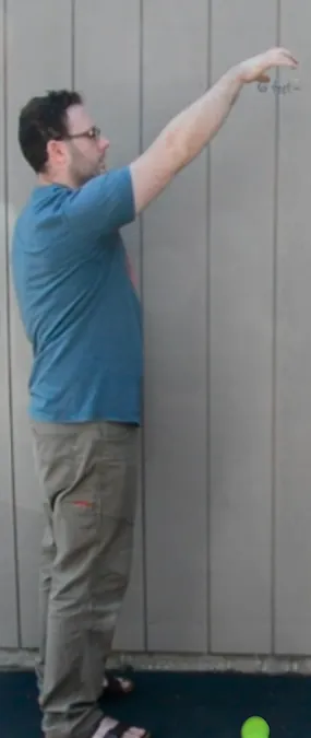 A man in a blue shirt and khaki pants stands sideways  against a gray paneled wall, arm extended upward, and has dropped a yellow tennis ball. The ball is on the ground.