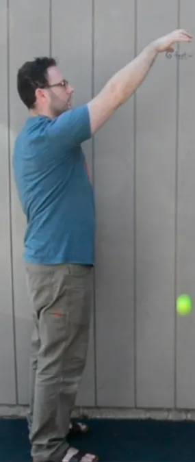 A man in a blue shirt and khaki pants stands sideways  against a gray paneled wall, arm extended upward, and has dropped a yellow tennis ball. The ball is approximately knee height of the man..
