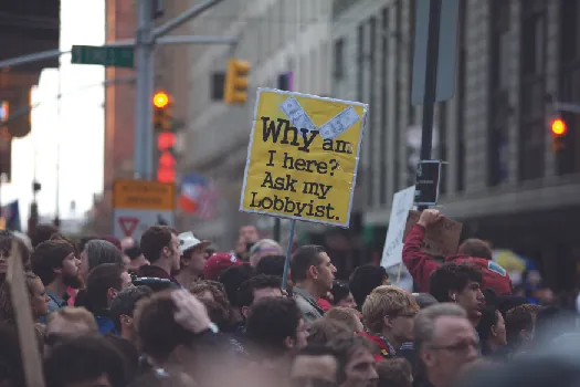 An image of a crowd of people, one of whom holds a sign that reads “Why am I here? Ask my lobbyist”.