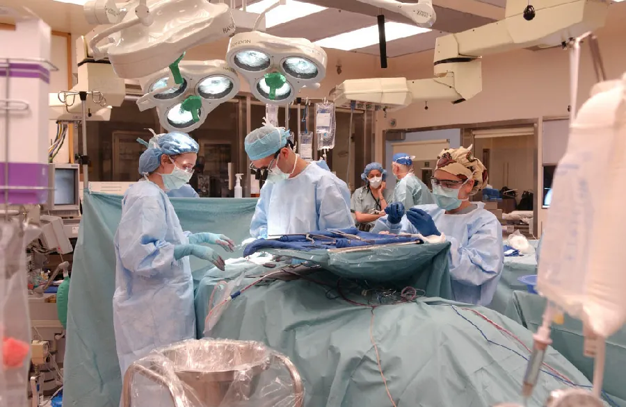 Photo of medical personnel performing surgery in room filled with medical equipment, sheets, lights, and staff assisting.