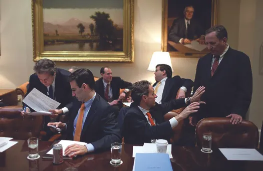 An image of seven people seated around a room. Some are talking to each other. Some are examining papers.