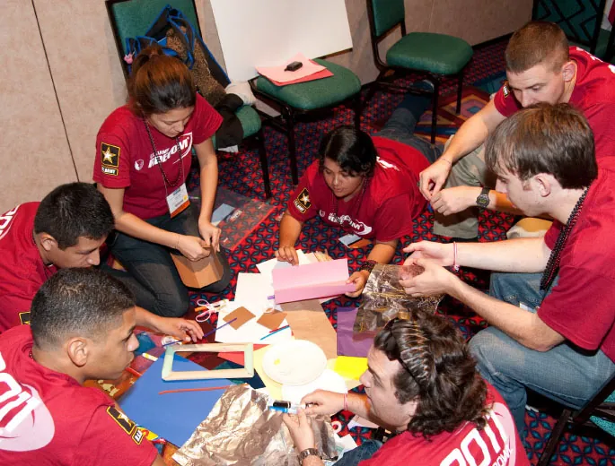 A group of ethnically diverse young adults, wearing matching red shirts, are seated on the floor around a variety of crafting materials, including colored paper, scissors, tape, and frames. They are working collaboratively on a project, engaging in a hands-on activity. They are in an indoor classroom environment, with chairs and tables around them.