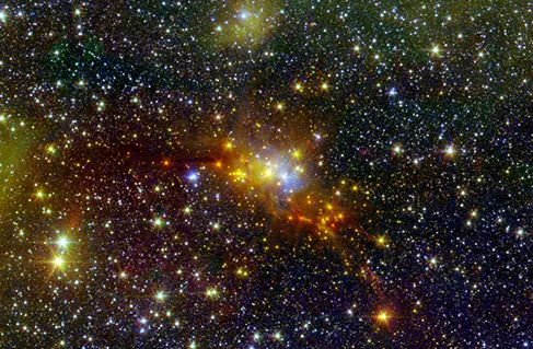 Una imagen de telescopio que muestra numerosas estrellas. Un cúmulo brillante en el centro tiene estrellas amarillas, naranjas y azules.
